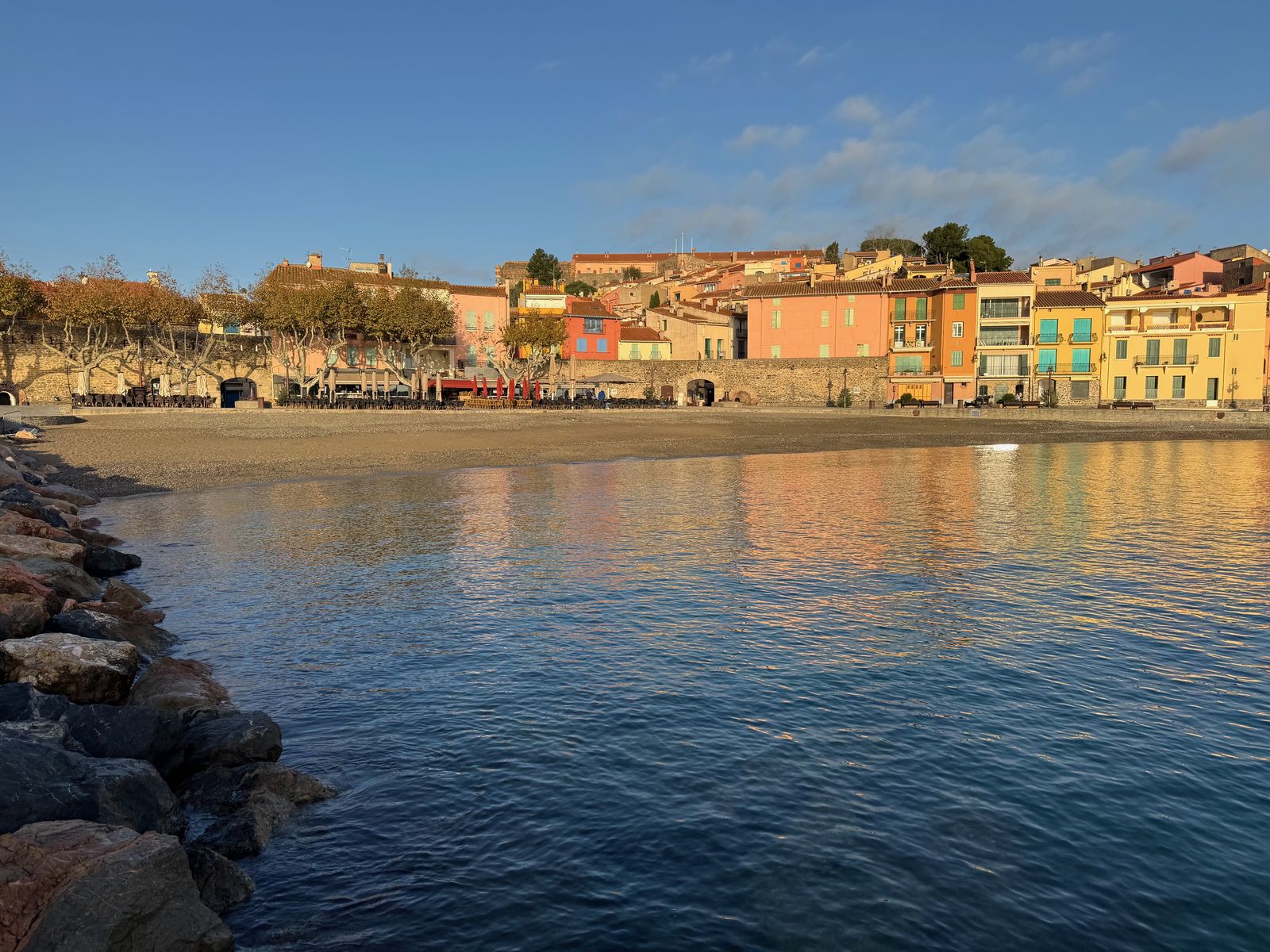 Le front de mer de Collioure au soleil rasant du matin, les terrasses encore repliées et la plage déserte